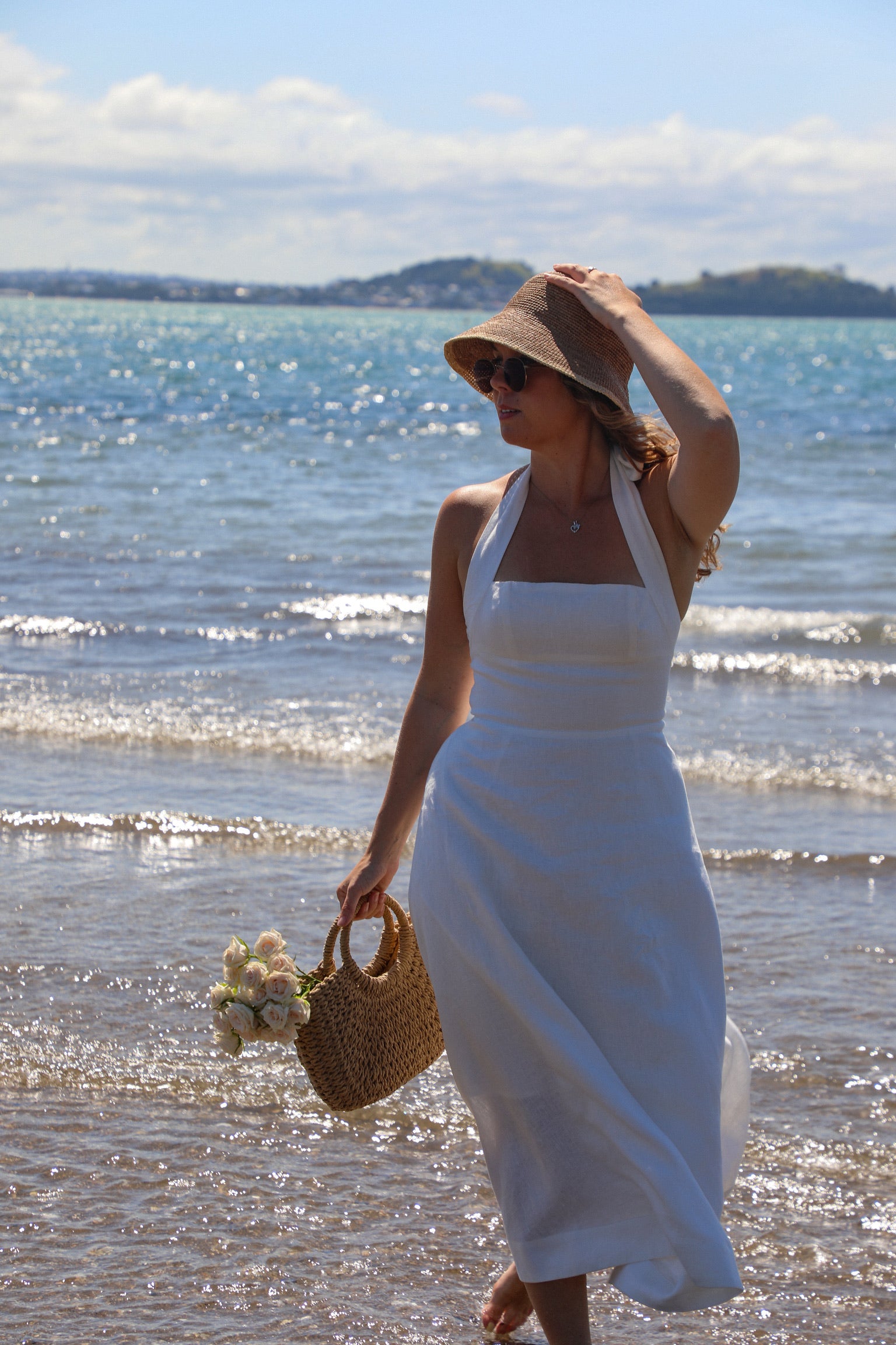 Woman in a white dress and straw hat standing on a beach with ocean waves in the background