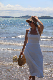 Woman in a white dress and straw hat standing on a beach with ocean waves in the background